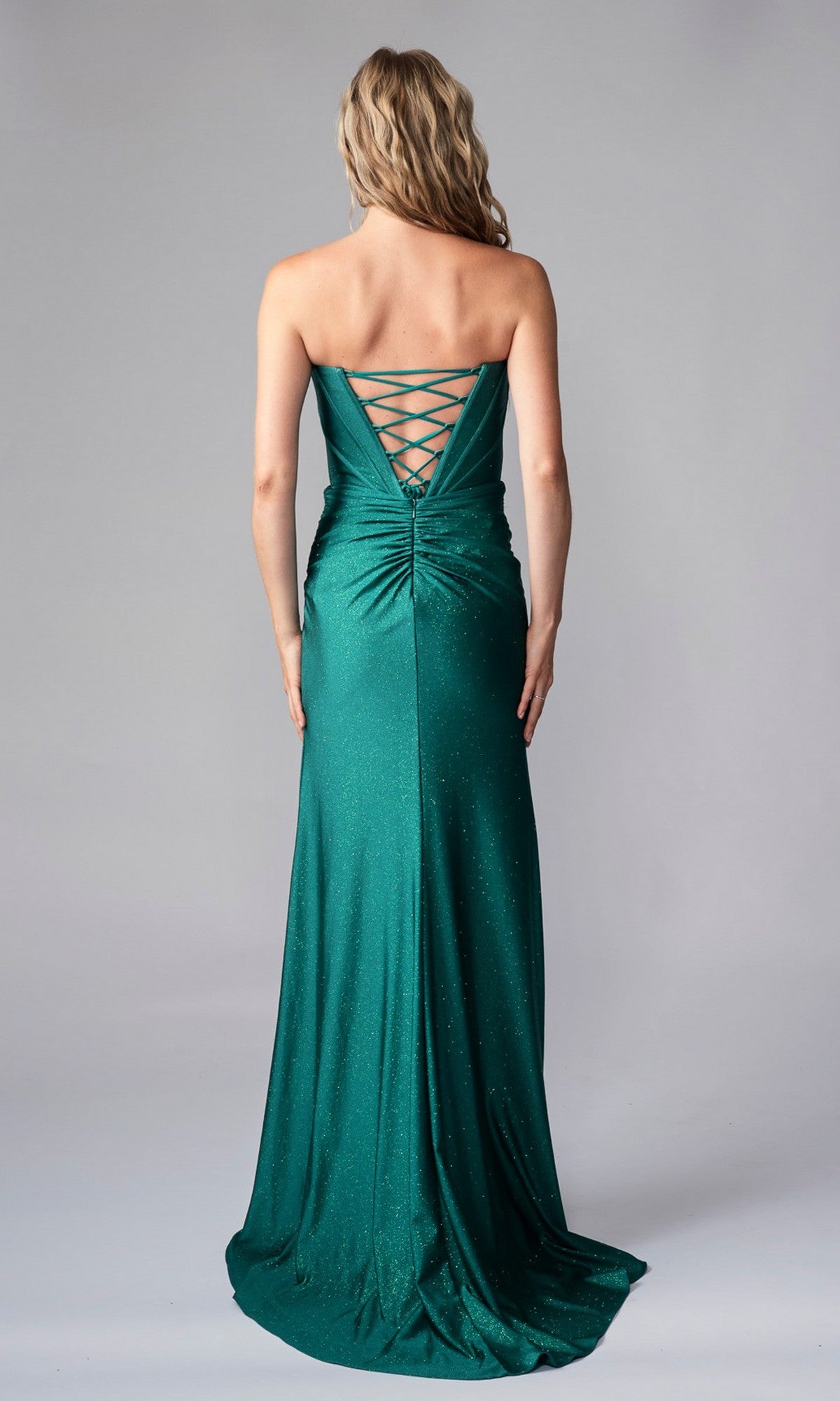 A woman faces away in a strapless emerald green Nina Canacci 1608 long formal dress, featuring a lace-up back and subtle ruching, set against a plain background.